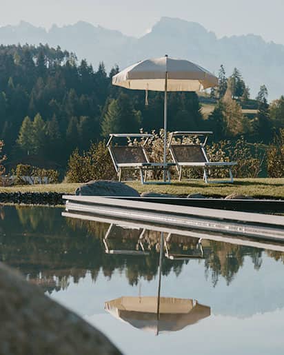 Liegestühle mit Sonnenschirmen am Naturpool des Naturhotels Pfösl in Deutschnofen, dahinter dichter Wald und ferne Berggipfel – ruhige Outdoor-Szene für Entspannung und Retreat.