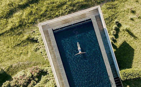 Luftaufnahme eines rechteckigen Outdoor-Pools inmitten einer grünen Wiese der Outdoor-Spa-Anlage des Naturhotels Pfösl; eine Person treibt entspannt auf dem Rücken im klaren Wasser.