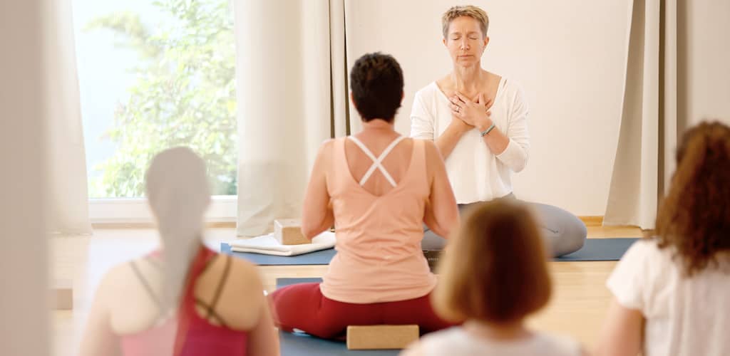 Yoga-Lehrerin Susanne Weisheit sitzt mit geschlossenen Augen und Händen auf dem Herzen vor Kursteilnehmerinnen in ruhiger Meditationshaltung